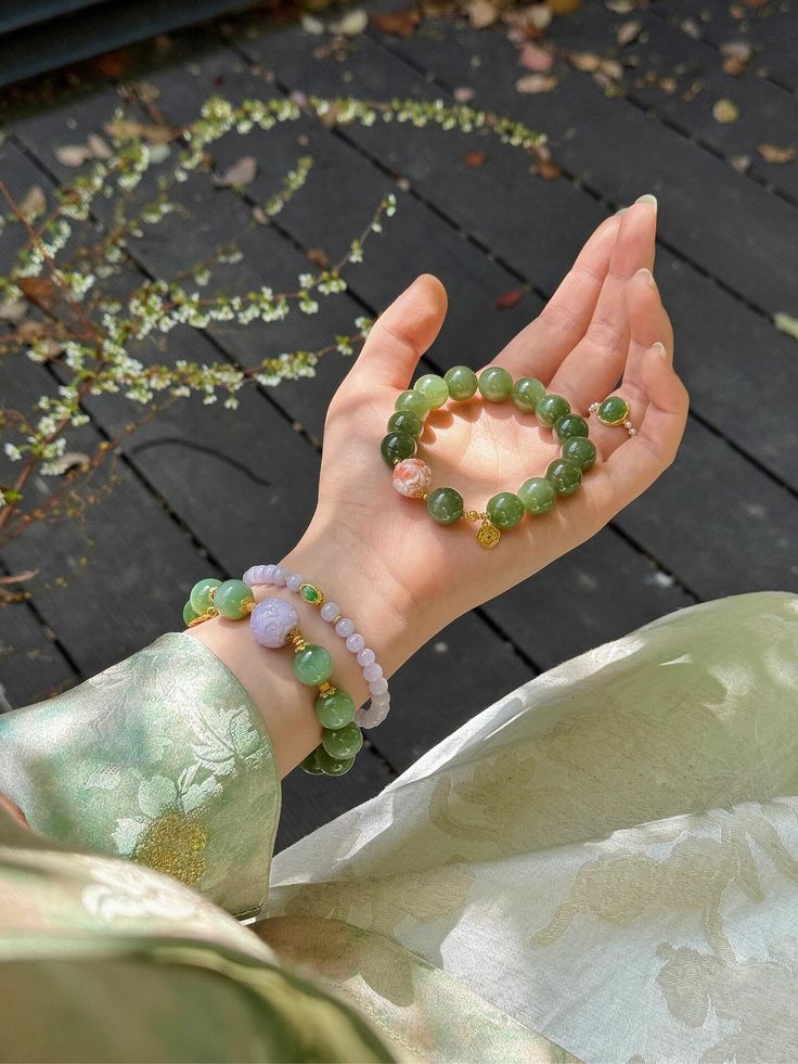 Bàn tay cầm vòng tay ngọc bích xanh và vòng ngọc tím lavender, charm vàng, trên nền gỗ sẫm